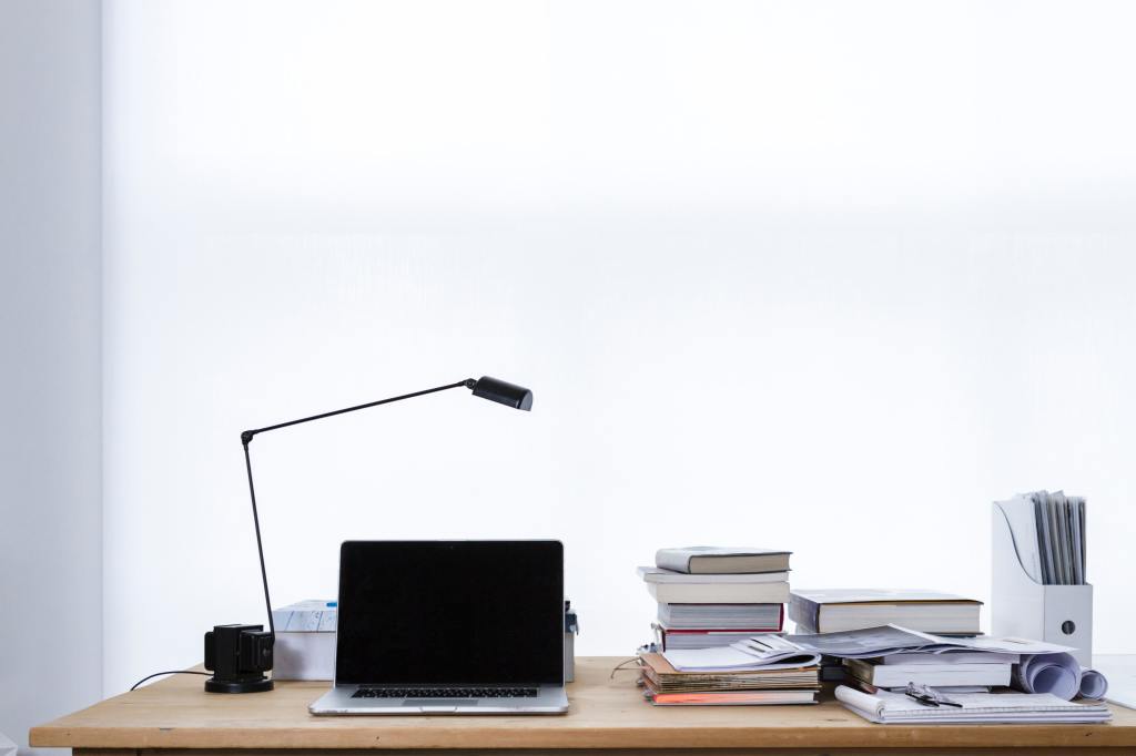 A desk with a desk lamp, laptop lots of books and file holder on it. 