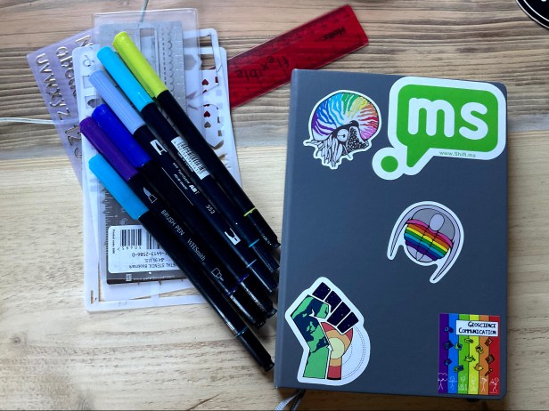 A grey leuchturm 1917 bullet journal with lots of different stickers. There is also a small pile of plastic stencils and brush pens.