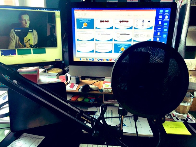 A photo of my recording set up. Boom mic at the front, two screens at at the back showing PowerPoint overview of slides and a second screen with a video of me recording the voice over.