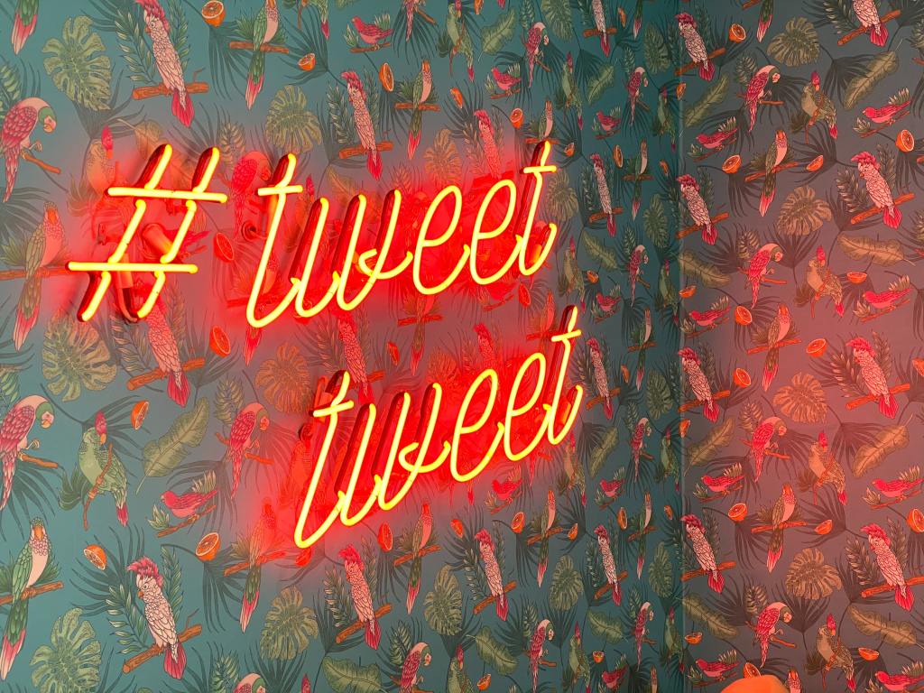 A pink-orange neon sign: # tweet tweet on a wall with tropical wallpaper. The wall paper is green and has images of branches with oranges and tropical birds on it. 