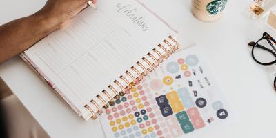 A person writing into a journal - the celebrations pages open. next to the journal is a sticker page.