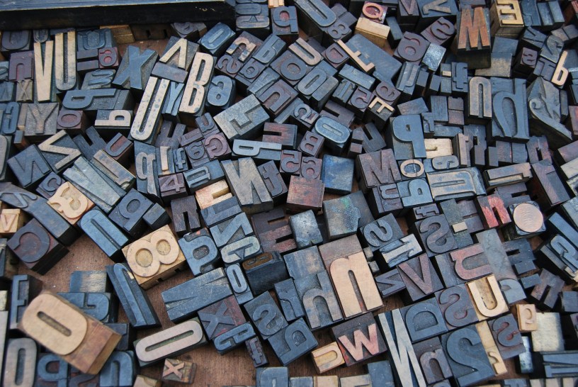 A collection of vintage letterpress printing blocks scattered across a wooden surface. The blocks are made of metal and wood, showing various letters and numbers in different sizes, arranged randomly. The blocks appear worn and weathered, with most appearing in blue-gray metallic tones, while some are in lighter wooden tones. The blocks are shown in reverse, as is typical for letterpress printing blocks, creating a jumbled, textural composition